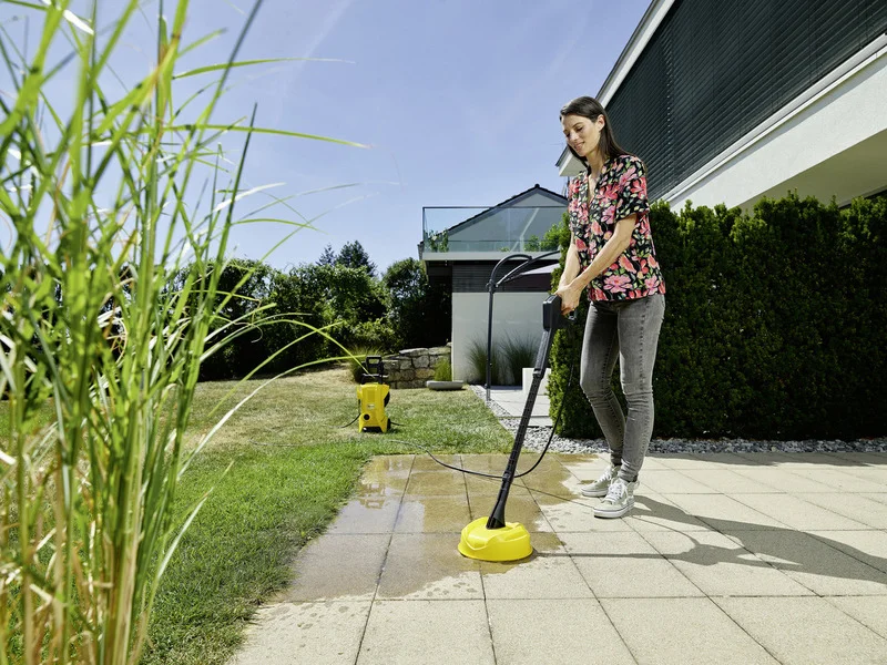 Person reinigt Terrasse mit Kärcher Hochdruckreiniger. Garten im Hintergrund, sonniges Wetter, moderne Architektur.