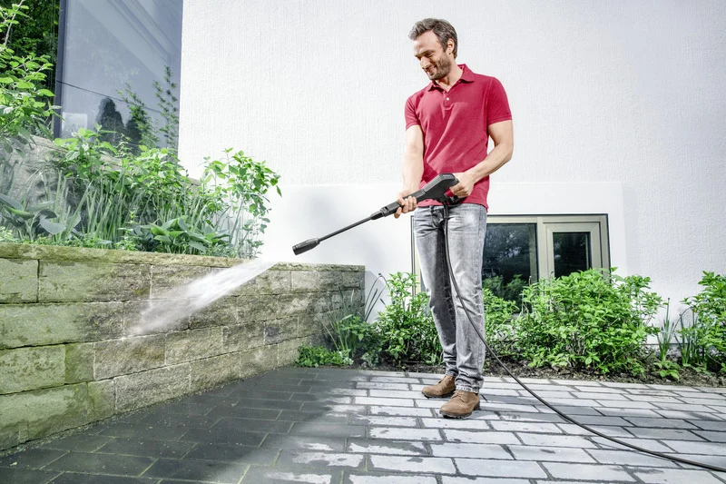 Mann reinigt mit Kärcher Hochdruckreiniger eine Steinmauer im Garten. Hintergrund: weiße Hauswand, Fenster und Pflanzen.