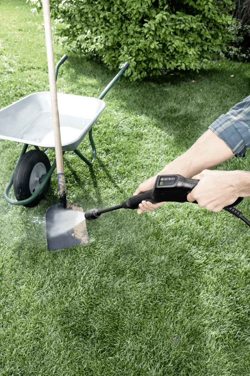 Person reinigt Schaufel mit Kärcher Hochdruckreiniger im Garten. Im Hintergrund steht eine Schubkarre.