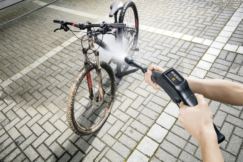 Person reinigt ein schlammiges Fahrrad mit einem Kärcher Hochdruckreiniger auf einem gepflasterten Boden im Freien.