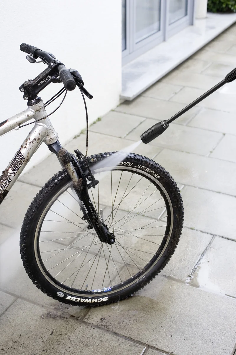 Ein schmutziges Fahrrad wird mit einem Kärcher Hochdruckreiniger auf einer gepflasterten Terrasse gereinigt.