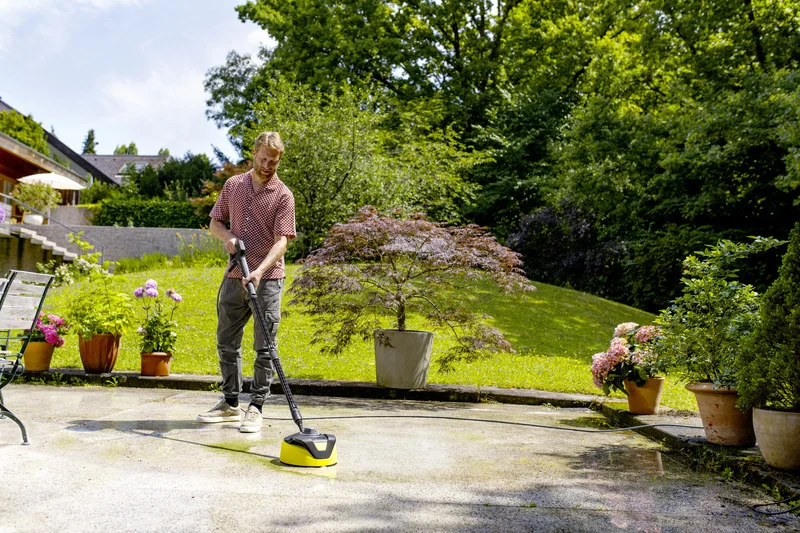 Mann reinigt Terrasse im Garten mit Kärcher Hochdruckreiniger, umgeben von blühenden Pflanzen und Bäumen.