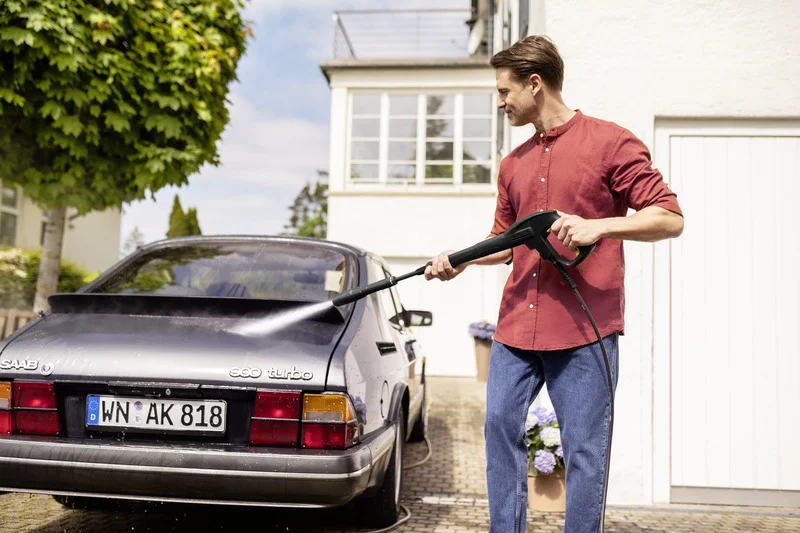 Mann reinigt schwarzen Saab 900 Turbo mit Kärcher Hochdruckreiniger im Freien vor einem Haus.
