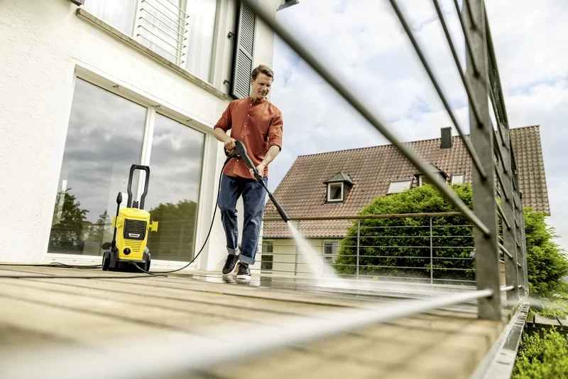 Person reinigt Terrasse mit Kärcher Hochdruckreiniger, im Hintergrund ein Haus mit Garten und blauer Himmel.