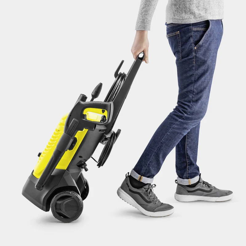 Person zieht einen gelben Kärcher Hochdruckreiniger auf zwei Rädern. Jeans und graue Schuhe sind sichtbar.