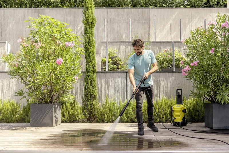 Mann reinigt Terrasse mit Kärcher Hochdruckreiniger, umgeben von blühenden Oleanderpflanzen und einer modernen Gartenmauer.
