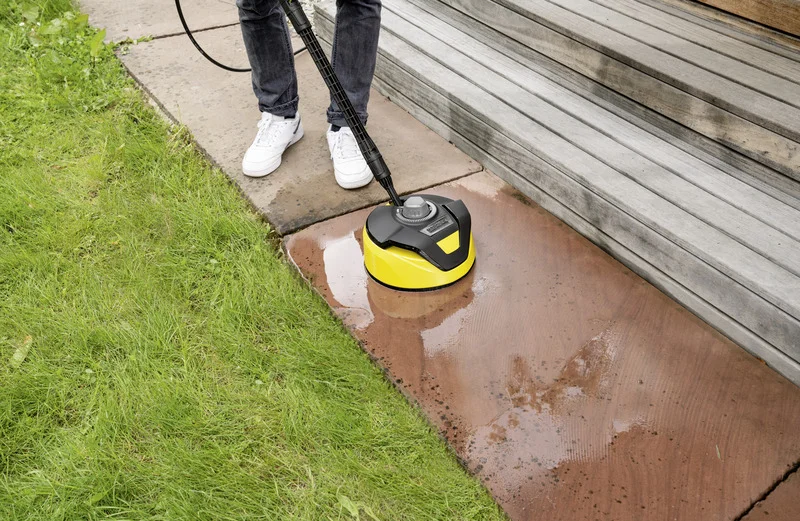 Person reinigt Terrasse mit Kärcher Flächenreiniger. Grüne Wiese und Holzstufen im Hintergrund. Weiße Schuhe und Jeans sichtbar.