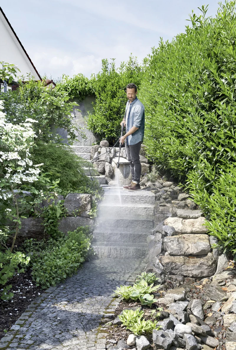 Person reinigt Steintreppe im Garten mit Kärcher Hochdruckreiniger, umgeben von grünen Büschen und Pflanzen.