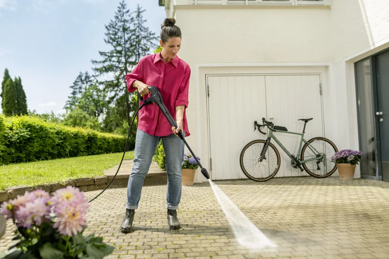 Person reinigt Terrasse mit Kärcher Hochdruckreiniger, Fahrrad lehnt an Garagentor, Blumen in Töpfen, sonniger Tag im Garten.