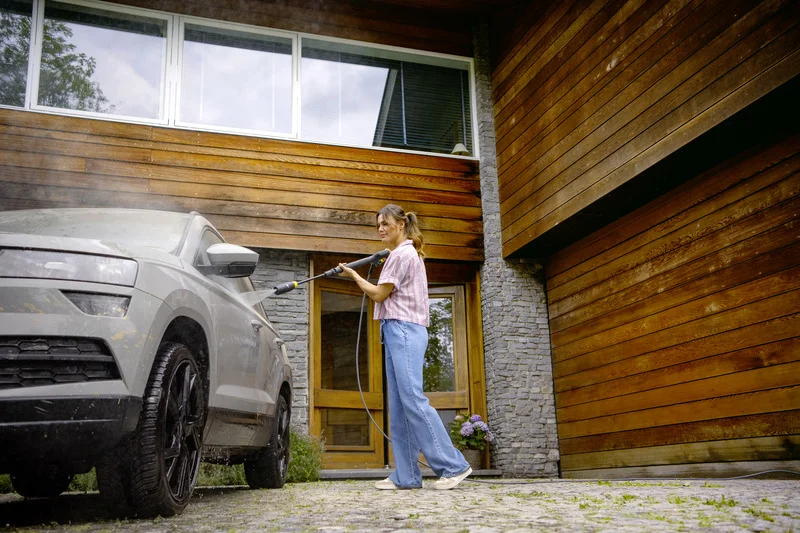 Person reinigt ein Auto mit einem Kärcher Hochdruckreiniger vor einem modernen Holzhaus.