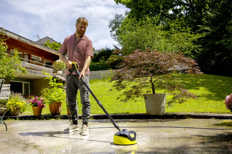 Person reinigt Terrasse im Garten mit Kärcher Hochdruckreiniger. Im Hintergrund sind Pflanzen und ein modernes Haus zu sehen.