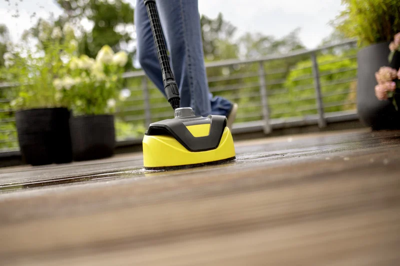 Kärcher Flächenreiniger reinigt Holzterrasse im Freien, Person in Jeans und Schuhe im Hintergrund. Pflanzen und Geländer sichtbar.
