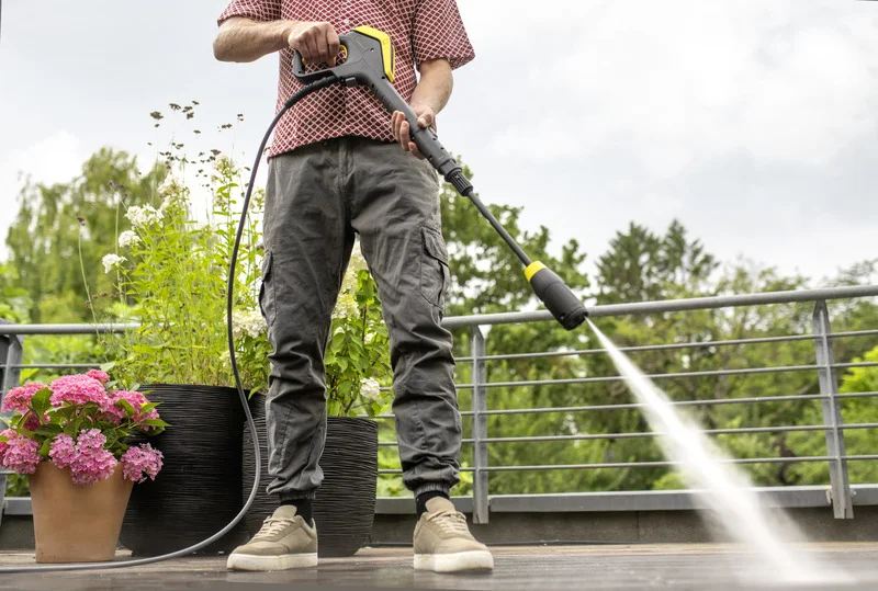 Person reinigt Terrasse mit Kärcher Hochdruckreiniger, umgeben von blühenden Pflanzen und Geländer im Freien.