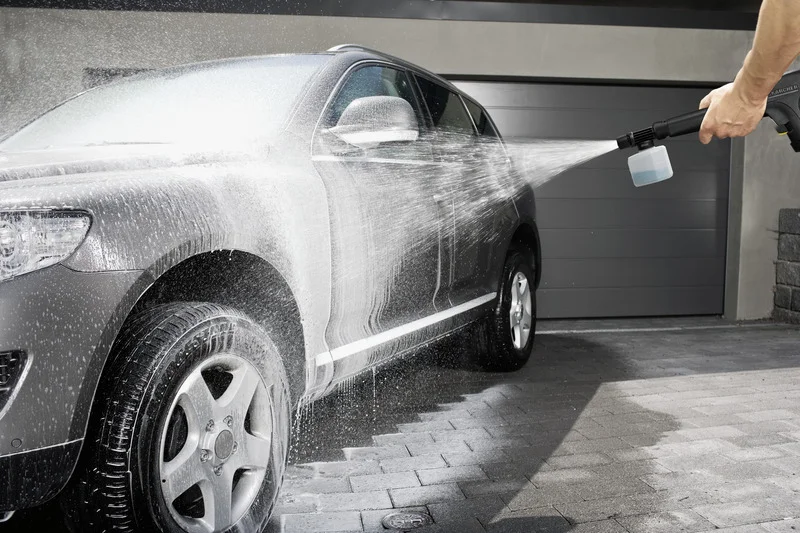 Silbernes Auto wird mit einem Kärcher Hochdruckreiniger gereinigt, Wasser spritzt auf der Einfahrt.