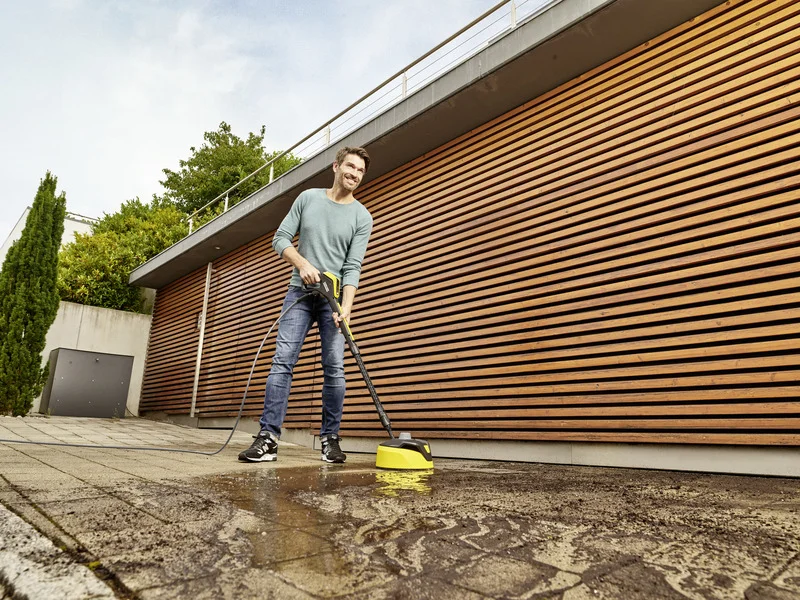 Person reinigt Terrasse mit Kärcher Bodenreiniger. Holzverkleidete Wand und grüne Pflanzen im Hintergrund. Outdoor-Szene.