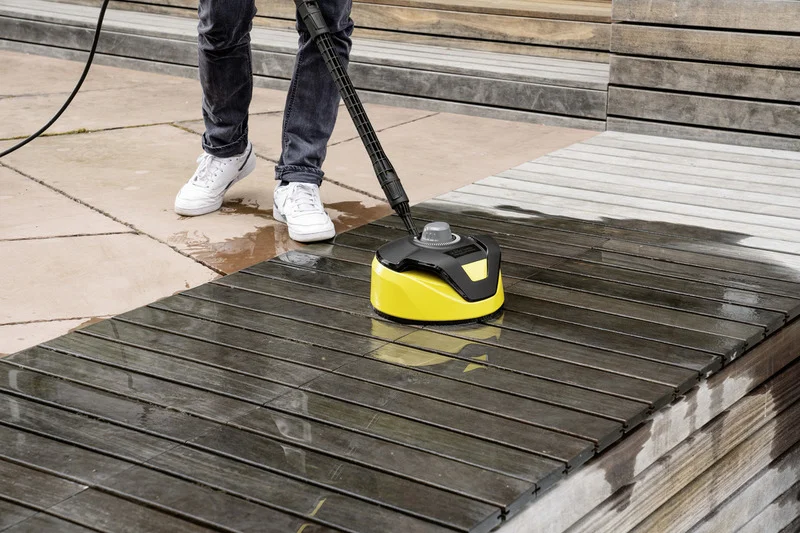 Person reinigt Holzterrasse mit Kärcher Flächenreiniger. Weiße Schuhe, Jeans. Terrasse halb gereinigt, halb verschmutzt.