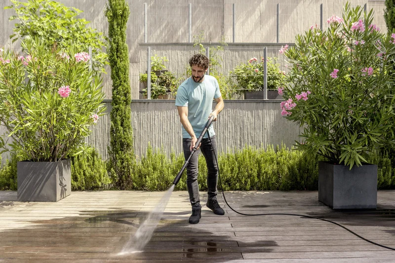 Mann reinigt Terrasse mit Kärcher Hochdruckreiniger, umgeben von blühenden Oleanderpflanzen und grüner Hecke im Garten.