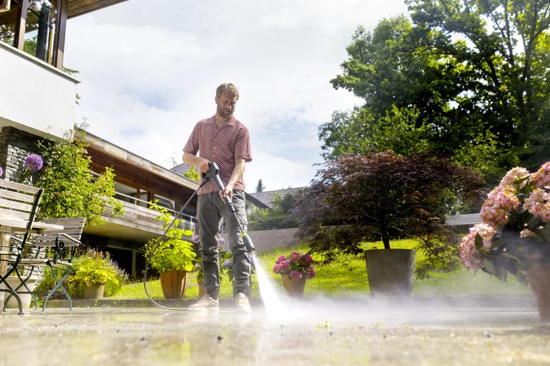 Person reinigt Terrasse mit Kärcher Hochdruckreiniger, umgeben von Pflanzen und Bäumen im Garten. Sonniger Tag, moderne Architektur im Hintergrund.