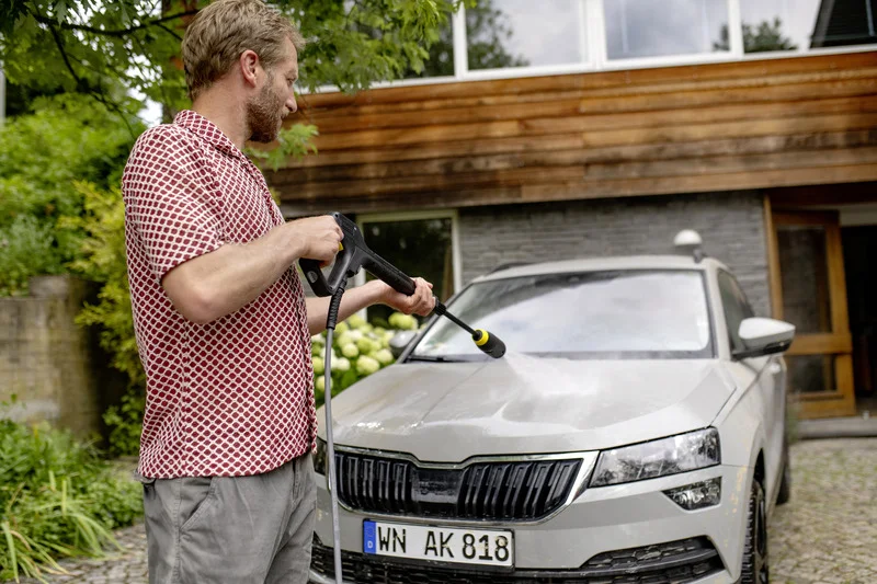 Mann reinigt graues Auto mit Kärcher Hochdruckreiniger im Freien vor einem Haus mit Holzfassade und Garten.