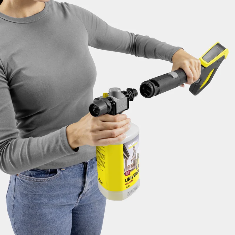 Person hält Kärcher Hochdruckreiniger und verbindet ihn mit einem gelben Reinigungsmittelbehälter. Graues Oberteil, blaue Jeans.
