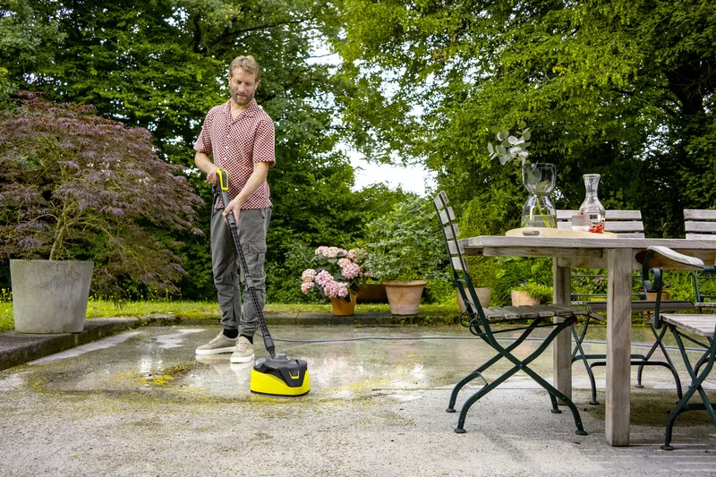 Person reinigt Terrasse mit Kärcher Hochdruckreiniger. Im Hintergrund Garten mit Pflanzen, Tisch mit Karaffe und Glas auf Terrasse.