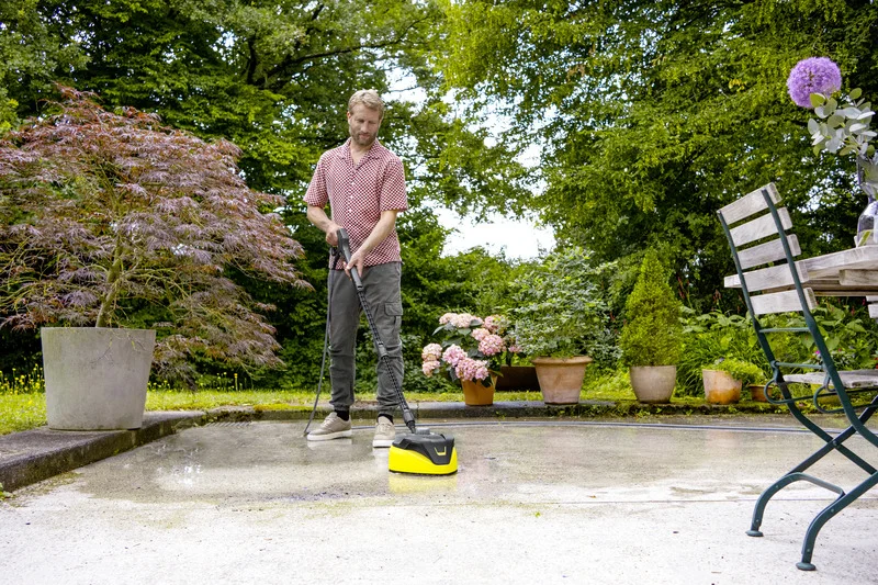 Person reinigt Terrasse mit Kärcher Hochdruckreiniger. Im Garten stehen Blumentöpfe und ein Tisch mit Stuhl.