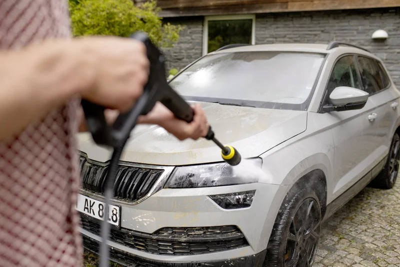 Person reinigt schmutziges Auto mit Kärcher Hochdruckreiniger im Freien. Vorderansicht des Autos mit Wasserstrahl auf Scheinwerfer.