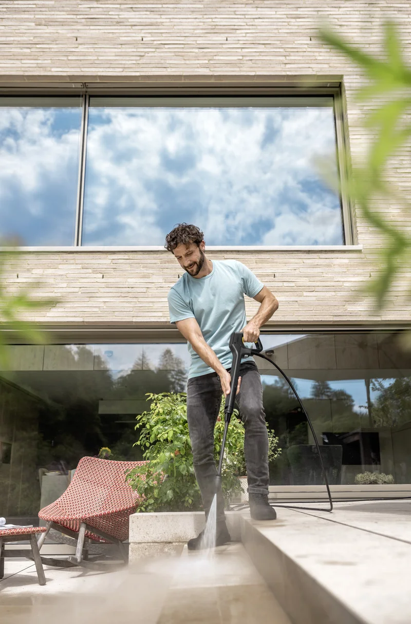 Mann reinigt Terrasse mit Kärcher Hochdruckreiniger, moderne Architektur im Hintergrund, Pflanzen und Gartenmöbel im Vordergrund.