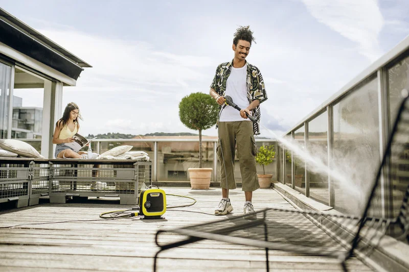 Person auf Terrasse nutzt Kärcher Hochdruckreiniger, während eine andere Person auf Sofa sitzt und liest. Sonniges Wetter, Pflanzen im Hintergrund.