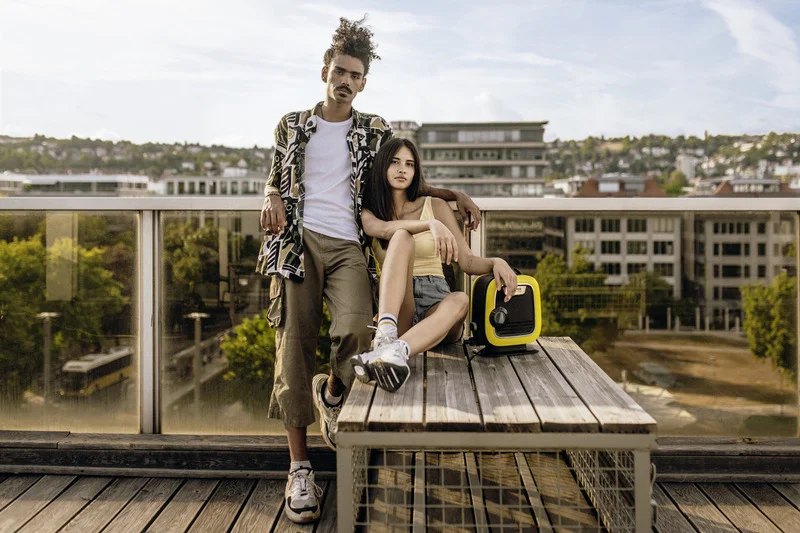 Zwei Personen sitzen auf einer Holzbank auf einer Dachterrasse, neben ihnen ein gelber Kärcher Lautsprecher. Im Hintergrund Stadtlandschaft.