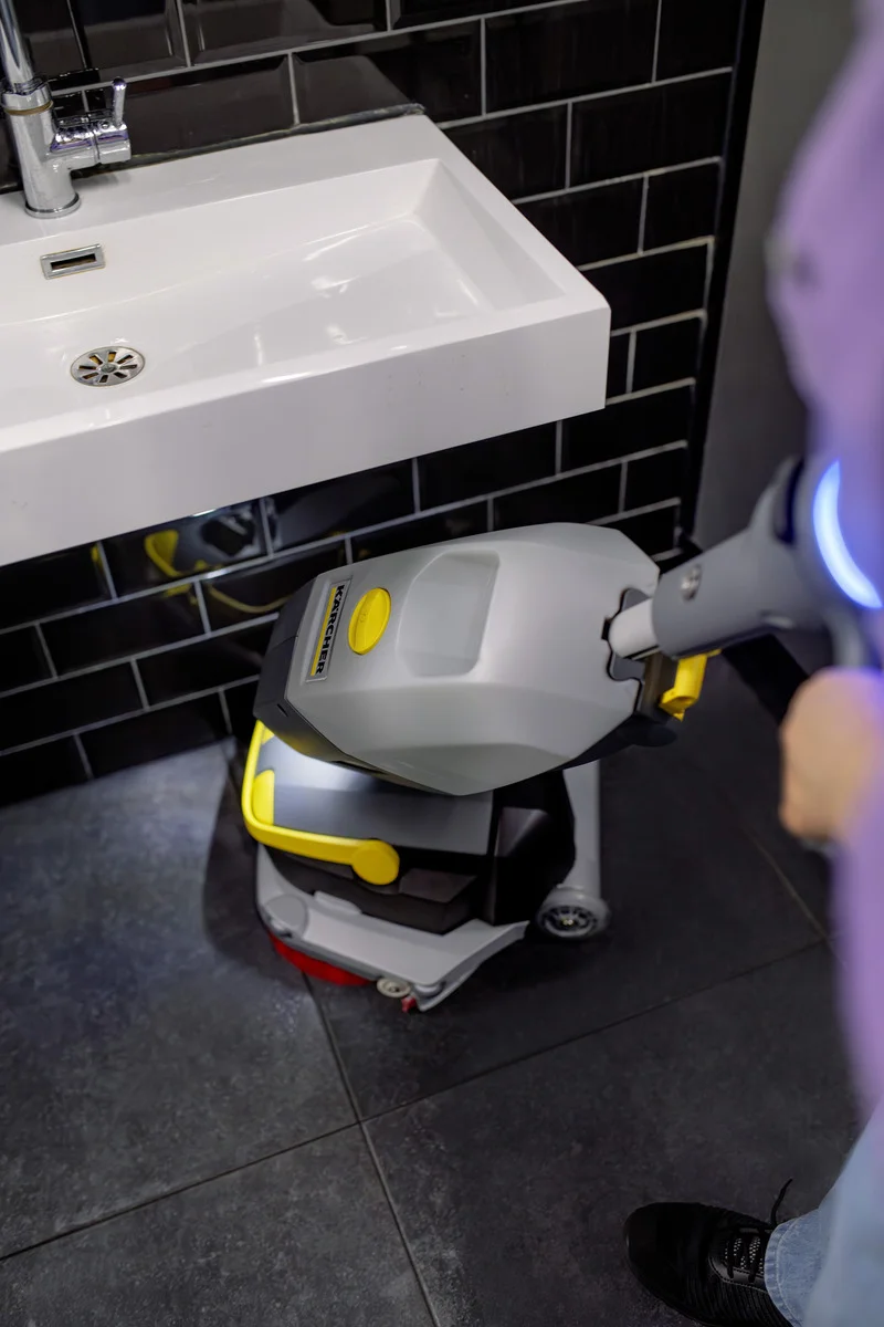Person reinigt Badezimmerboden mit Kärcher Bodenreiniger neben weißem Waschbecken und schwarzen Fliesenwänden.