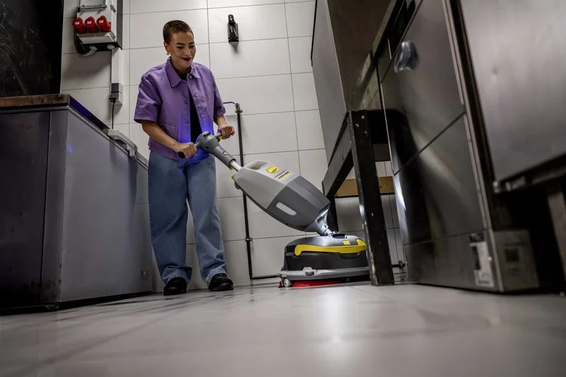 Mann reinigt Küchenboden mit Kärcher Bodenreinigungsmaschine in einem industriellen Umfeld. Er trägt ein lila Hemd und blaue Hose.