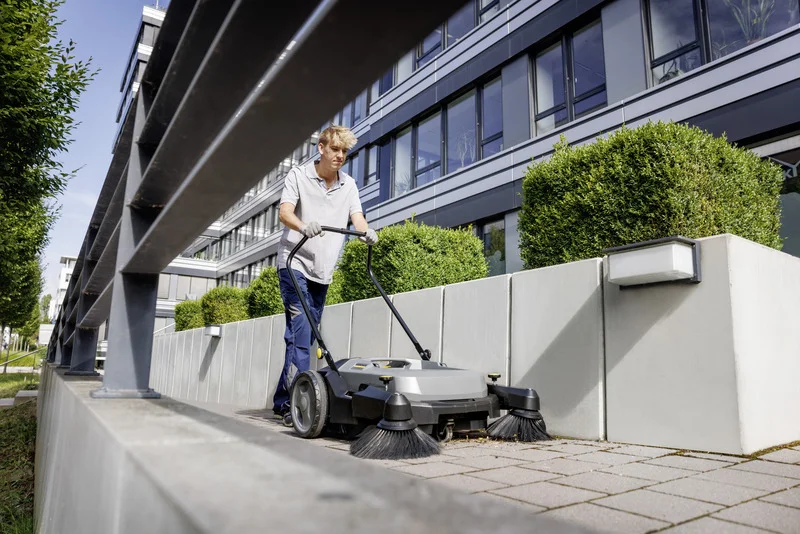Person reinigt Gehweg mit Kärcher Kehrmaschine, umgeben von modernen Gebäuden und grünen Büschen im Freien.