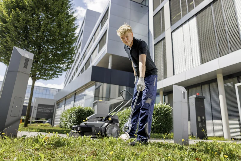 Person in Arbeitskleidung sammelt Müll mit Greifzange auf Grünfläche vor einem modernen Bürogebäude.