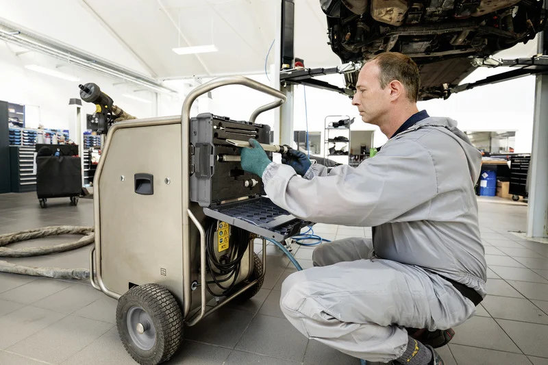 Mechaniker in Werkstatt bedient Kärcher Trockeneisstrahlgerät, Auto auf Hebebühne im Hintergrund.