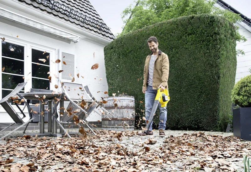 Person nutzt Kärcher Laubbläser auf einer Terrasse, um Herbstblätter zu entfernen. Im Hintergrund sind Gartenmöbel und eine Hecke zu sehen.