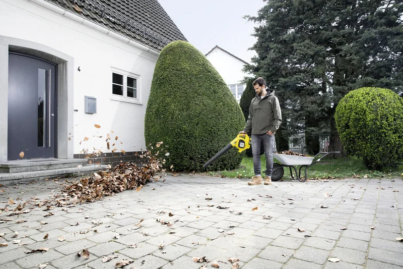 Person nutzt Kärcher Laubbläser, um Blätter auf gepflastertem Weg vor einem Haus zu sammeln. Schubkarre mit Blättern daneben.