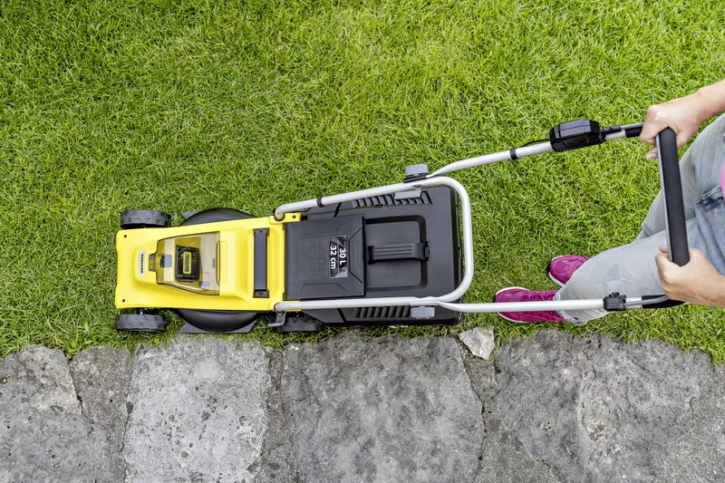 Person schiebt einen gelben Kärcher Rasenmäher über grünen Rasen neben einem Steinweg. Ansicht von oben.