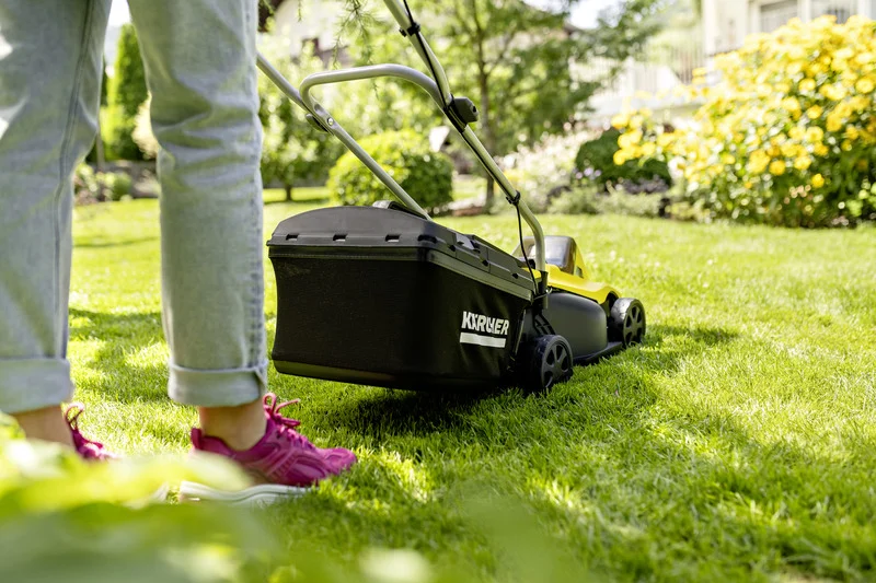 Person mit rosa Schuhen schiebt einen Kärcher Rasenmäher auf grünem Rasen im Garten. Im Hintergrund blühende Pflanzen.