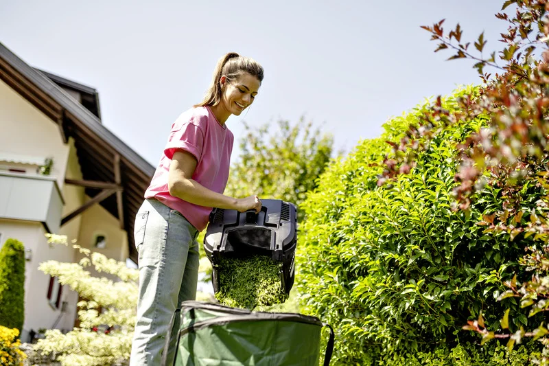 Frau leert Grasfangkorb in grünen Sack im Garten. Im Hintergrund ein Haus und grüne Hecke. Sonniger Tag.
