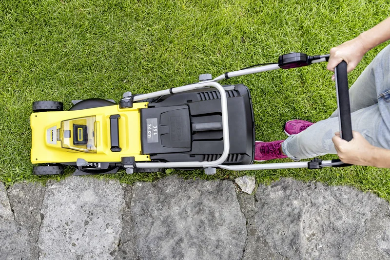 Person schiebt Kärcher Rasenmäher über Gras neben Steinweg, von oben gesehen. Rote Schuhe und Jeans sichtbar.