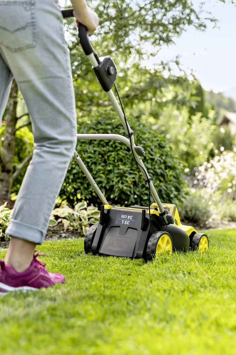 Person schiebt Kärcher Rasenmäher über grünen Rasen im Garten. Trägt Jeans und rote Turnschuhe. Hintergrund: Bäume und Sträucher.