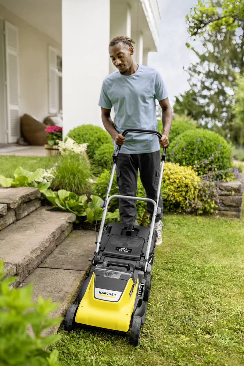 Person mäht Rasen mit einem Kärcher Rasenmäher im Garten vor einem Haus mit Pflanzen und Blumen.