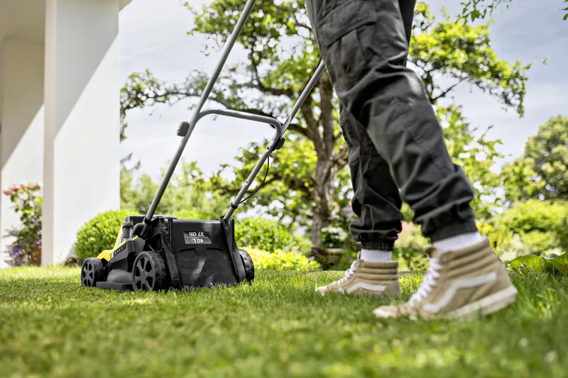 Person mäht Rasen mit einem Kärcher Rasenmäher im Garten. Hintergrund zeigt Bäume und Pflanzen.