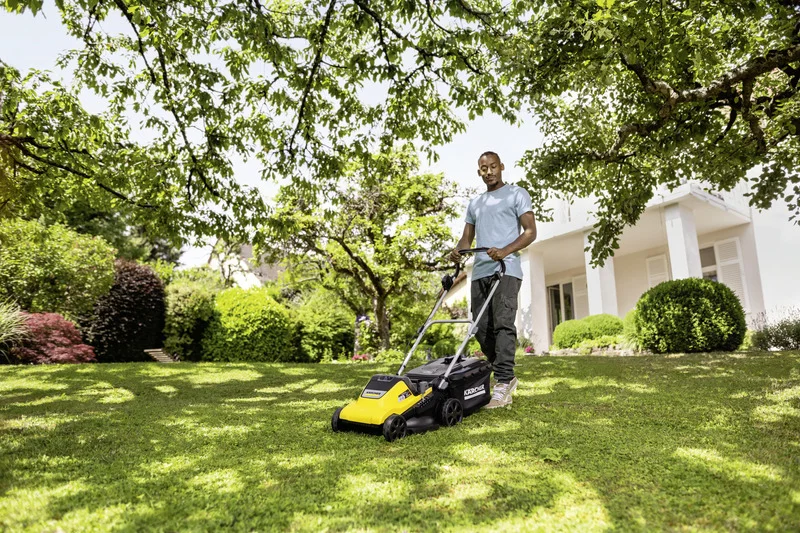 Person mäht Rasen mit einem Kärcher Rasenmäher im Garten vor einem modernen Haus, umgeben von Bäumen und Sträuchern.