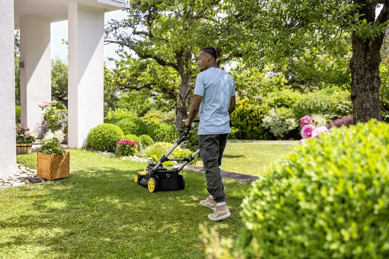 Person mäht Rasen mit einem Kärcher Rasenmäher in einem gepflegten Garten, umgeben von Büschen und Blumen.