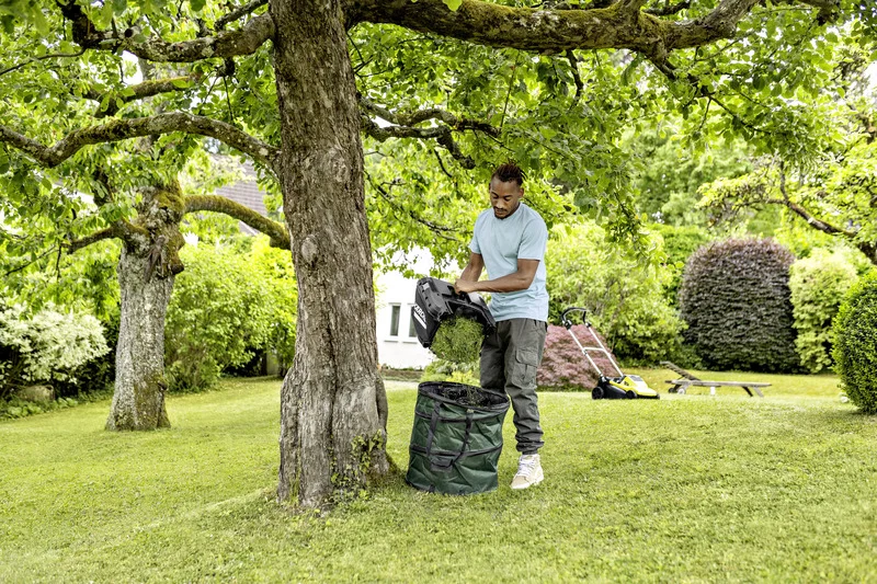 Mann leert Grasfangkorb eines Kärcher Rasenmähers in einen grünen Sack unter einem Baum im Garten.