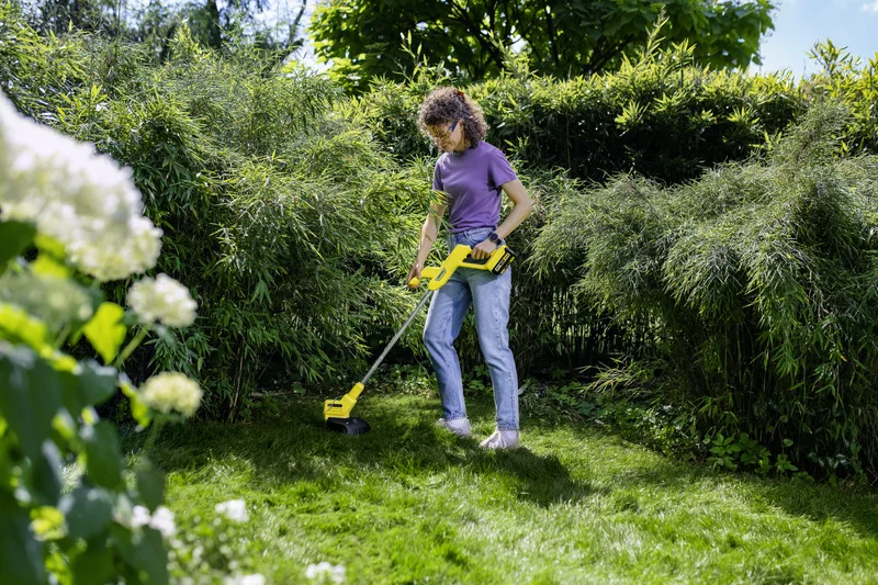 Frau in lila Shirt und Jeans benutzt Kärcher Rasentrimmer im Garten, umgeben von grünen Büschen und weißen Blumen.