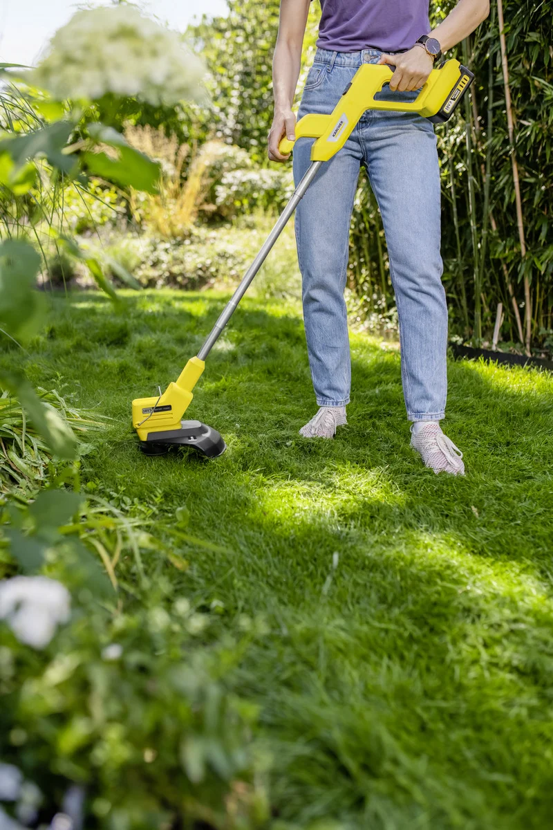 Person mit lila Shirt und blauer Jeans benutzt einen gelben Kärcher Rasentrimmer im grünen Garten.