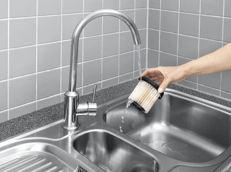 Hand reinigt Filter unter fließendem Wasser in Edelstahlspüle. Hintergrund aus grauen Fliesen.